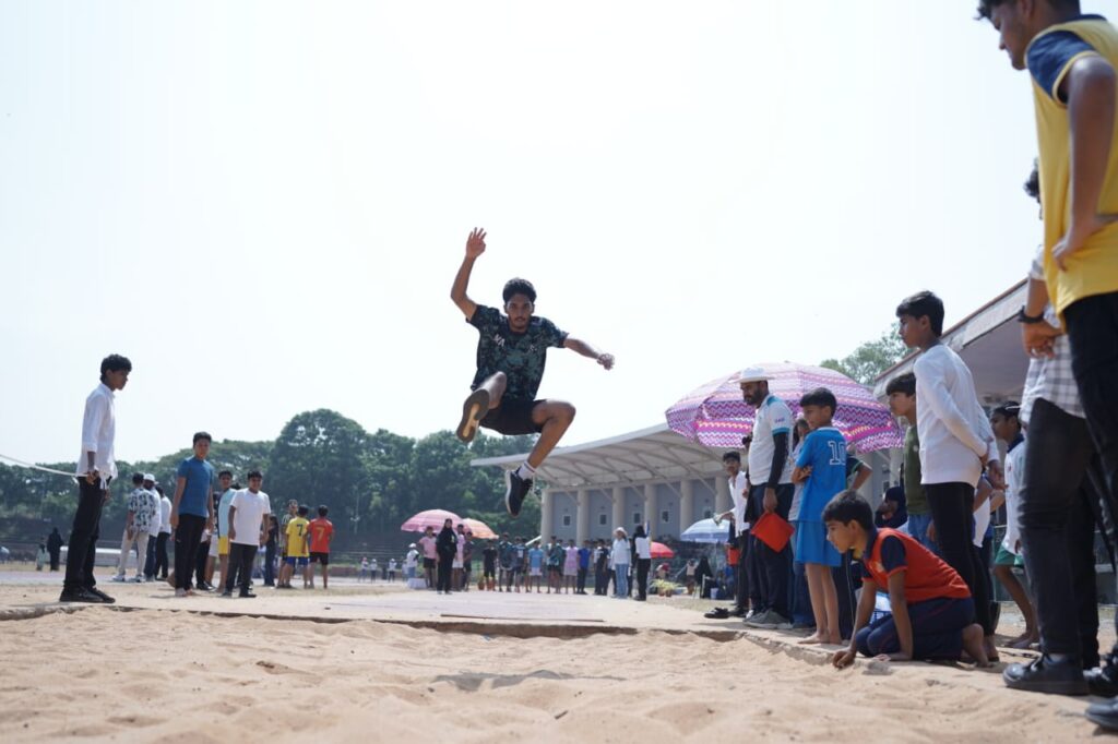 Prestige school student performing a long jump during a sports event on campus, with classmates and teachers watching and cheering.