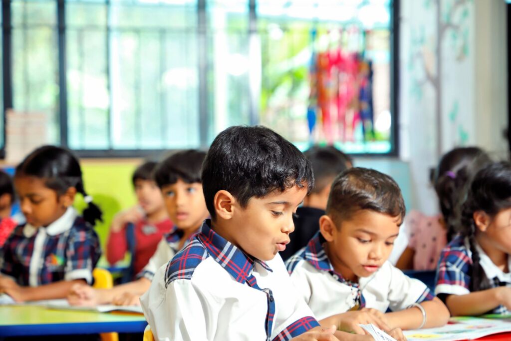Young school children sitting at desks in a brightly lit classroom, wearing uniforms and reading or writing in notebooks during a class activity.